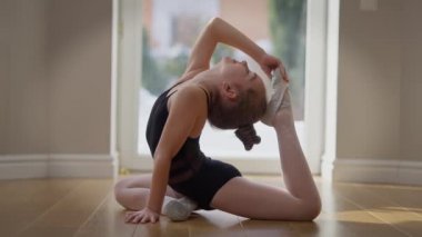 Side view flexible teen gymnast bending holding foot at head indoors at home. Portrait of motivated confident cute Caucasian girl training working out. Sport and lifestyle concept. Slow motion