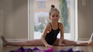 Wide shot teenage girl sitting on twine bending forward in slow motion. Portrait of confident concentrated Caucasian adolescent gymnast exercising at home indoors. Motivation and sport concept