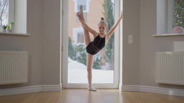 Wide shot flexible talented teenage gymnast stretching one leg up looking at camera standing indoors. Portrait of skilled Caucasian teen girl posing training in slow motion. Grace and flexibility