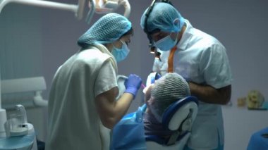 Focused female Caucasian dental assistant helping professional Middle Eastern dentist removing saliva from patient mouth. Concentrated young nurse assisting expert doctor treating man in dental chair