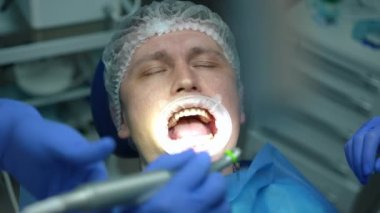 Doktor ve hemşire olarak dişçi koltuğunda gözleri kapalı bir hastanın yüzü matkap ve tükürük püskürtücüyle belirdi. Ağır çekimde diş kliniğinde tedavi gören beyaz adamın yakın plan portresi.