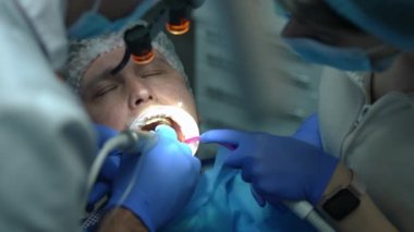 Headshot of Caucasian man with mouth retractor opening eyes as dentist leaving with drill and assistant removing saliva with sucking tube. Close-up portrait of patient undergoing treatment in hospital