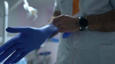 Yakın çekimde tanınmayan bir dişçi tek kullanımlık eldivenlerini giyer ve hastanede ağır çekimde durur. Profesyonel Ortadoğulu kendine güvenen doktor klinikteki hasta ziyaretine hazırlanıyor