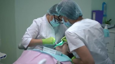 Focused female orthodontist curing ill tooth of patient in dental chair with assistant removing saliva with ejector. Professional concentrated Caucasian women working in medical clinic indoors