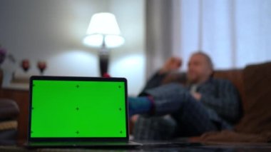Close-up chromakey laptop with blurred Caucasian man sitting on couch at background reading book. Unrecognizable relaxed guy in pajamas enjoying hobby with green screen device at front