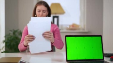 Sağda Chromakey dizüstü bilgisayarı ve arka planda evrakları analiz eden bulanık genç bir kadın var. Ev ofisinde çalışan, kendine güvenen, Kafkasyalı, serbest çalışan bir kadın. Yaşam tarzı ve serbest çalışma