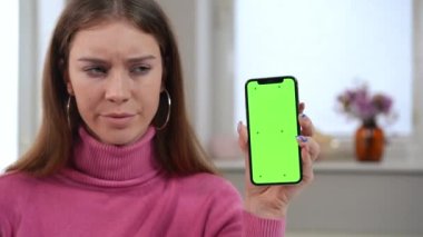 Close-up chromakey smartphone in hand of dissatisfied young woman shaking head. Portrait of unsatisfied Caucasian millennial lady posing with green screen phone. Social media web page mockup