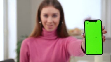 Arka planda gülümseyen bulanık, yeşil ekran akıllı telefon. İnce, beyaz, kendine güveni tam, mutlu bir kadın. Kapalı alanda kromakey telefon reklamcılığı uygulaması ile poz veriyor.
