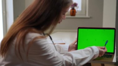 Unrecognizable blurred doctor pointing at green screen on laptop talking advertising web page mockup. Young slim Caucasian woman promoting treatment online site in hospital indoors
