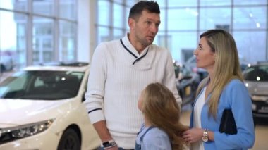 Unsure Caucasian man choosing new car in dealership with family talking looking around. Portrait of handsome successful husband father discussing vehicle purchase with wife and daughter in slow motion
