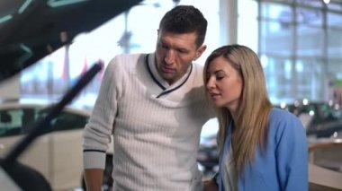 Positive woman talking with concentrated man examining open car trunk in car dealership. Beautiful Caucasian wife discussing automobile purchase with successful handsome husband in showroom