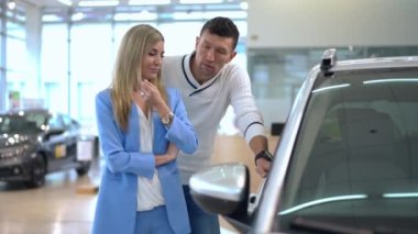 Positive beautiful woman and handsome man talking choosing new automobile in car dealership. Medium shot portrait of Caucasian successful couple making decision standing in showroom indoors