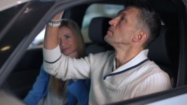 Side view concentrated successful man on drivers seat examining car with blurred smiling woman talking at background on passenger seat. Rich Caucasian couple choosing new automobile in dealership