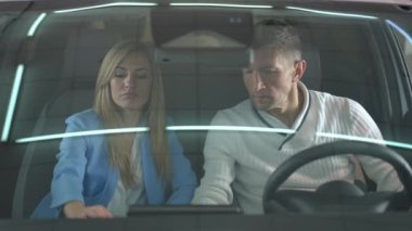 Car windshield with couple sitting in salon making decision buying new vehicle. Front view portrait of concentrated successful Caucasian man and woman talking choosing automobile in car dealership