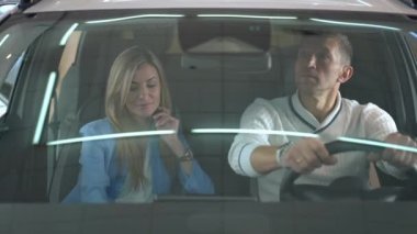 Thoughtful man and woman sitting in car with cute girl entering sitting on back seat. Shooting through windshield of successful Caucasian family choosing new automobile in dealership smiling talking