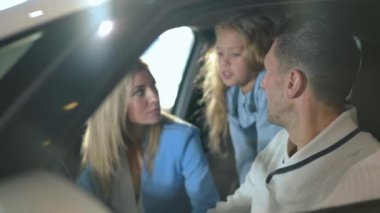Side view man smiling listening blurred woman and girl talking at background sitting in new automobile in car dealership. Positive confident Caucasian father husband choosing vehicle with family