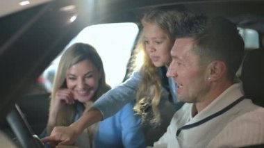 Happy father showing car dashboard to curios cute daughter with blurred mother smiling at background. Side view portrait of confident Caucasian family choosing new automobile in dealership together