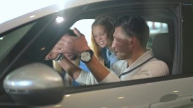 Side view cheerful man laughing talking with woman and girl sitting in car in showroom. Portrait of positive Caucasian joyful husband father choosing new vehicle with family in car dealership