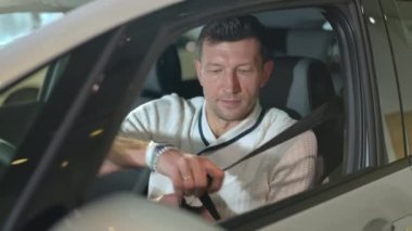 Handsome man fastening seat belt sitting on drivers seat in new luxurious automobile in car dealership. Portrait of satisfied Caucasian buyer choosing new vehicle in car showroom indoors