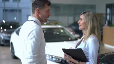 Yan görüş, araba galerisindeki beyaz otomobili işaret eden beyaz araba satıcısı ve alıcısı hakkında olumlu. Kendine güveni tam bir kadın, başarılı bir adama araba satıyor.