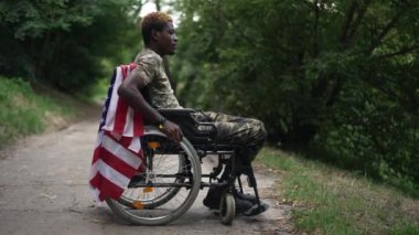 Side view thoughtful depressed African American young soldier in wheelchair with USA flag looking away thinking. Frustrated handsome man in summer park outdoors. Patriotism and disability