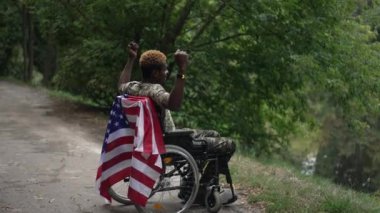 Soldier in wheelchair gesturing strength in slow motion admiring lake in forest. Side view wide shot African American young man with US flag enjoying leisure outdoors. Individuality and disability