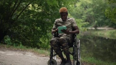 Smiling African American man in wheelchair surfing social media on tablet sitting on lake shore in summer park. Wide shot portrait of positive confident relaxed soldier enjoying leisure outdoors