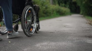 Unrecognizable Caucasian woman pushing wheelchair with African American man in summer park. Young girlfriend taking care of military boyfriend outdoors. Support and disability concept