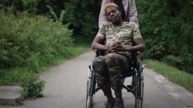 Tekerlekli sandalyedeki kendinden emin Afro-Amerikan adam ve tanınmayan beyaz bir kadın hareket yardım aracı kullanıyor. Parkta sevgilisiyle birlikte pozitifliğin Dolly portresi.