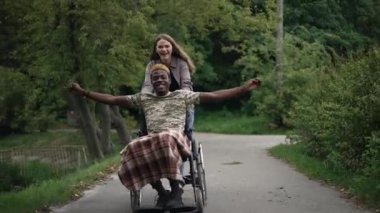 Joyful African American man in wheelchair stretching hands smiling as cheerful Caucasian woman pushing device. Portrait of happy positive interracial couple having fun in summer park travelling