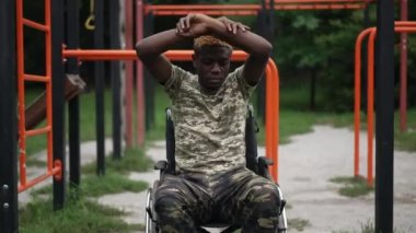 Arka planda spor ekipmanlarıyla dışarıda ısınan tekerlekli sandalyedeki Afro-Amerikalı adama kendinden emin bir şekilde. Yaz parkındaki cesur asker eğitiminin portresini büyüt. Spor ve sakatlık