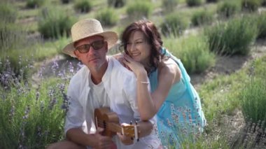 Loving woman smiling holding shoulder of talented man playing ukulele in slow motion in sunshine. Portrait of happy Caucasian adult girlfriend dating with boyfriend on lavender field on summer day