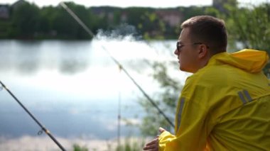 Side view Caucasian fisherman smoking sitting on the right with fishing rods on river bank. Young concentrated relaxed man in sunglasses and yellow raincoat fishing on summer morning in park