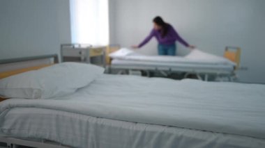 Hastane koğuşunda beyaz, temiz bir yatak. Arka planda yatağını yapan bulanık bir genç kadın var. Modern, rahat bir klinikte, tanınmayan, Kafkasyalı hasta. Sağlık ve Hizmet