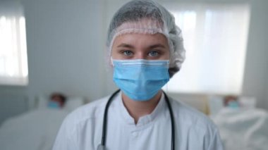 Close-up woman in coronavirus uniform looking at camera leaving hospital ward with ill patients at background. Young Caucasian doctor in face mask walking away. Covid-19 pandemic outbreak concept