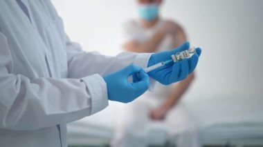 Tanımlanamayan doktor şırıngayı aşıyla dolduruyor arka planda bekleyen bulanık bir hasta gibi yatakta oturuyor. Eldivenli ve üniformalı profesyonel hemşire Covid-19 aşısı için hastanede ilaç hazırlıyor.