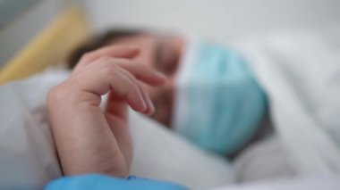 Hasta, yorgun, beyaz, Covid-19 maskeli, hemşire avuç içiyle yatakta yatan adamın eli. Tıbbi klinikteki hastane koğuşunda kadın bakıcısı olan yorgun erkek hasta.
