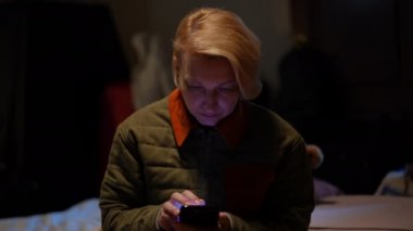 Devastated woman scrolling smartphone touchscreen sitting in bomb shelter during Russian bombardment. Portrait of hopeless sad beautiful Ukrainian lady reading news online hiding in basement floor.