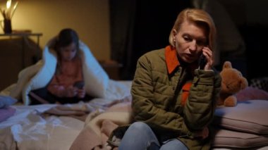 Worried woman talking on phone hiding from blurred girl playing on smartphone at background in bomb shelter. Portrait of anxious Ukrainian mother hiding with daughter on air-raid alert warning