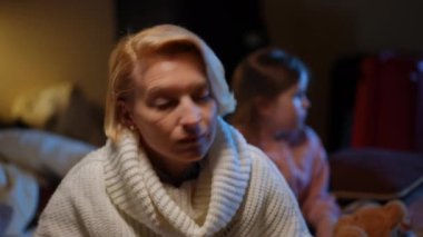 Anxious mother hugging scared daughter as air raid alert lamp flashing at background. Frightened Ukrainian woman and girl embracing hearing explosions sound hiding in bomb shelter