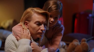 Close-up portrait of fatigue Ukrainian mother hugging frightened daughter with red air raid alert lamp flashing at background in bomb shelter. Tired exhausted woman and girl hiding embracing