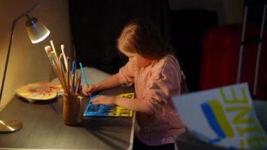 Side view focused sad girl drawing Help Ukrainian children placard sitting at table in bomb shelter. Concentrated cute stressed child painting indoors. Lost childhood and military invasion concept