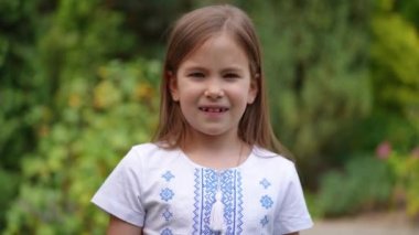 Brunette charming Ukrainian girl in national shirt drawing colors of flag on cheek looking away talking. Portrait of pretty child posing in summer garden outdoors. Childhood and Ukraine concept