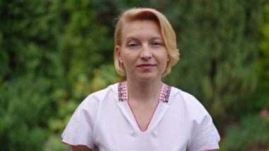 Front view adult woman in traditional Ukrainian shirt with embroidery looking at camera smiling. Proud beautiful lady posing in green garden outdoors in slow motion. Patriotism concept