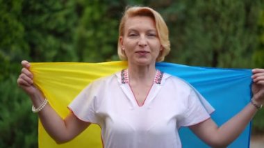 Front view portrait of confident brave woman posing with Ukrainian flag in slow motion. Medium shot of positive lady in embroidered shirt looking at camera standing outdoors in summer garden park