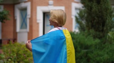 Woman with Ukrainian flag turning in slow motion looking at camera smiling standing in backyard garden. Portrait of confident lady posing outdoors on summer day wearing traditional embroidered shirt