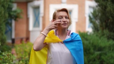 Medium shot portrait of proud Ukrainian woman wrapped in national flag drawing blue and yellow on cheeks smiling looking at camera. Patriotic lady posing in backyard garden outdoors