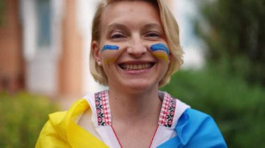 Headshot portrait of positive woman in embroidered shirt and Ukrainian flag looking at camera smiling in slow motion. Close-up front view proud lady posing outdoors on backyard