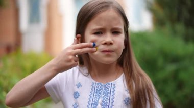 Cute Ukrainian girl looking at camera drawing yellow and blue on cheek standing in garden in traditional clothing with embroidery. Confident child posing in slow motion with serious facial expression