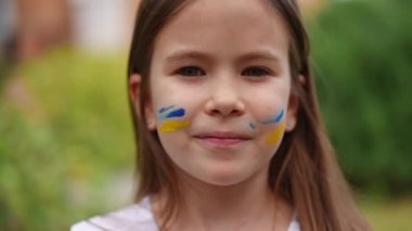 Pretty girl with brown eyes and face painting looking at camera smiling. Ukrainian child with flag colors on cheeks posing in slow motion outdoors. Pride and childhood concept